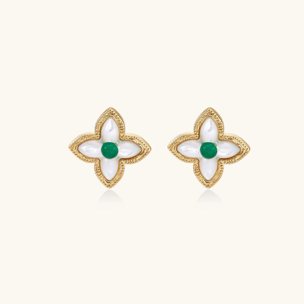 Gold quatrefoil stud earrings with white mother-of-pearl petals and emerald green gemstone center on beige background.