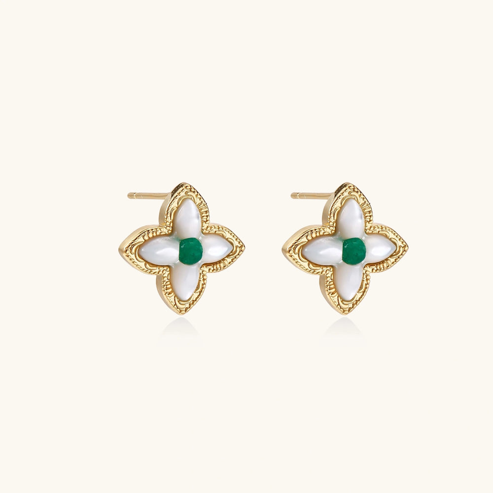Gold quatrefoil stud earrings with white mother-of-pearl petals and emerald green gemstone center on beige background.